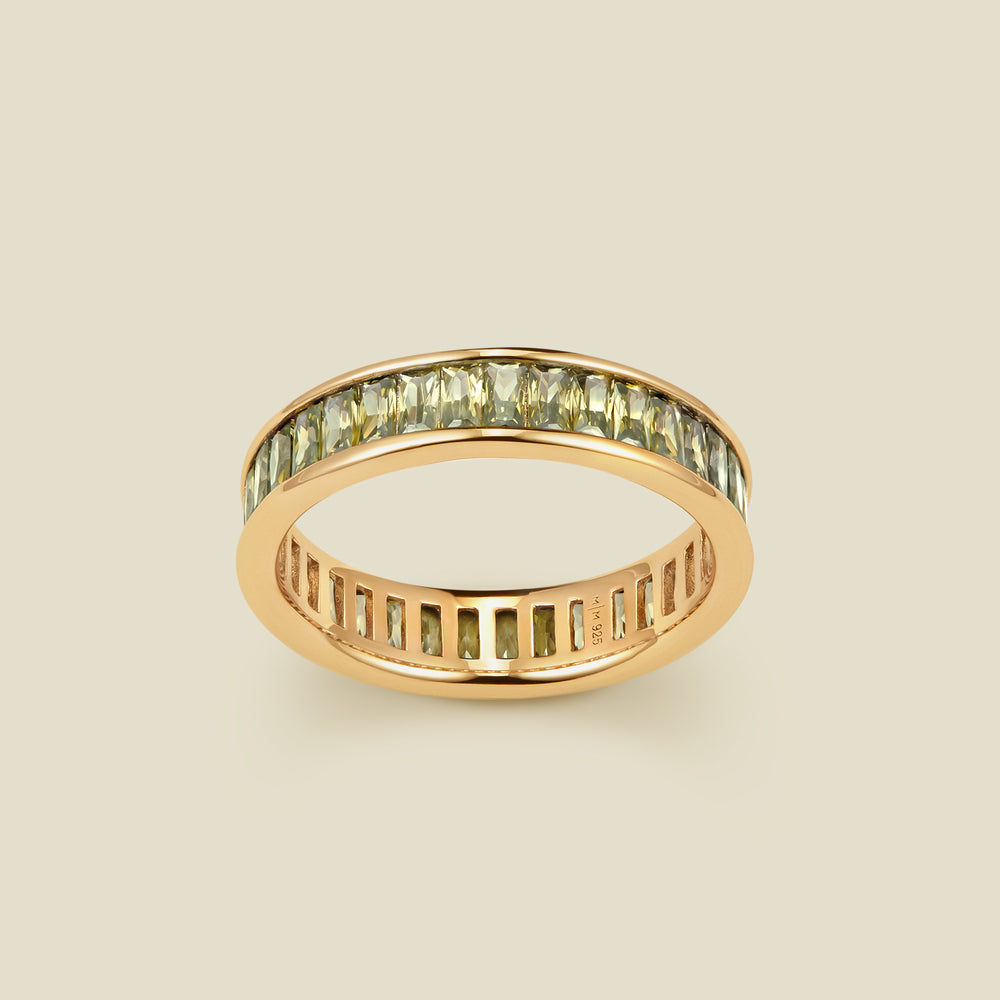 A gold eternity ring featuring a continuous channel setting of baguette-cut light green gemstones, shown on a solid light beige background.