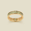 A gold eternity ring featuring a continuous channel setting of baguette-cut light green gemstones, shown on a solid light beige background.