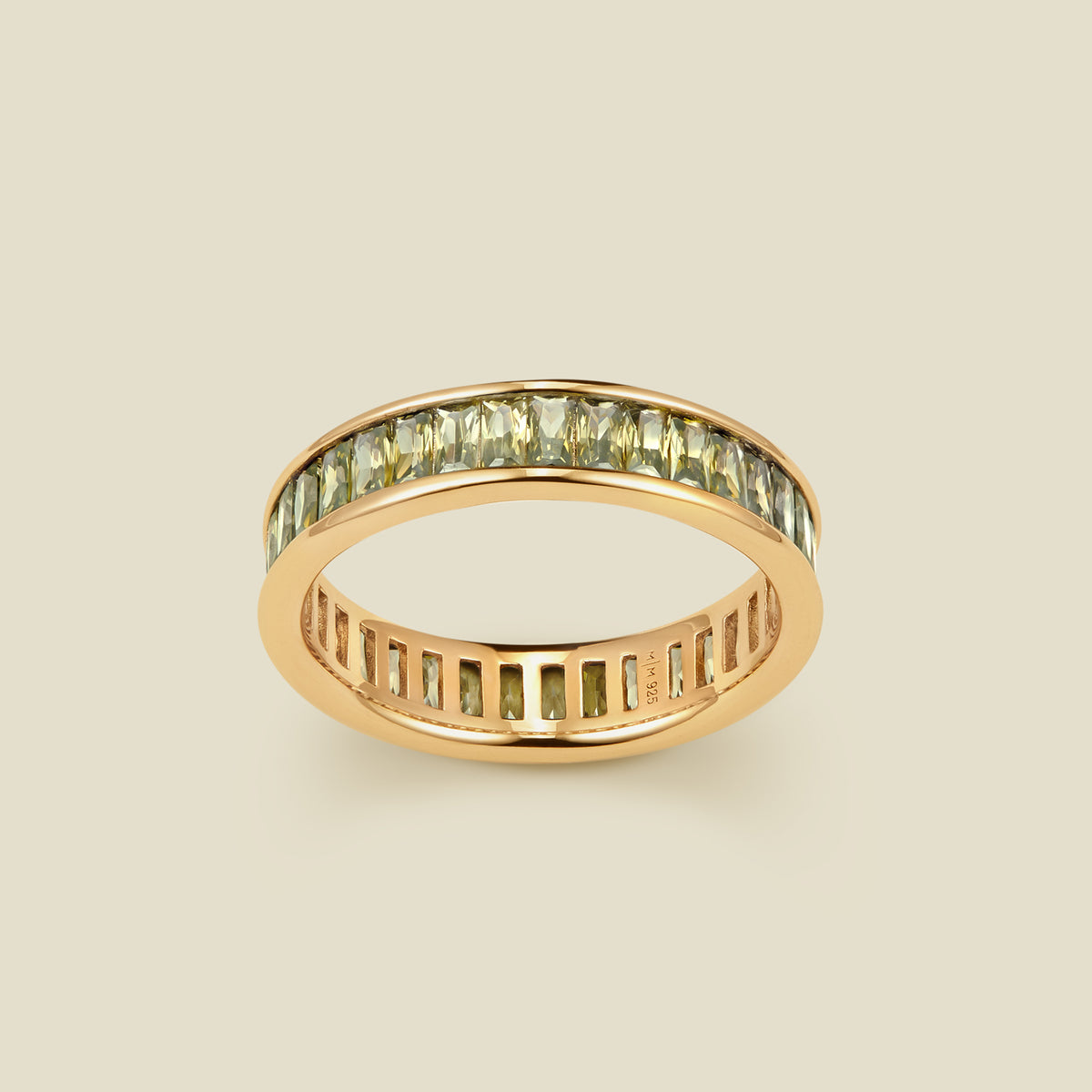 A gold eternity ring featuring a continuous channel setting of baguette-cut light green gemstones, shown on a solid light beige background.