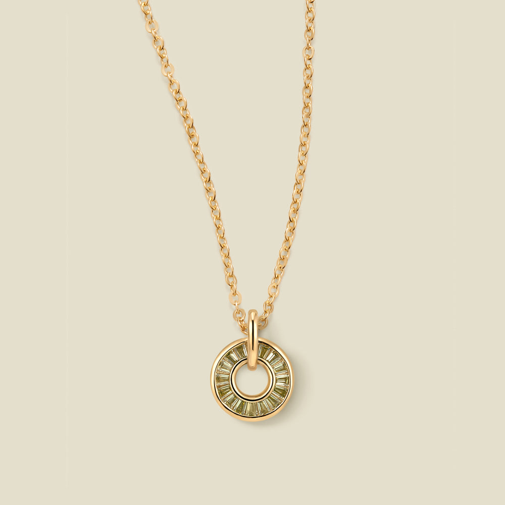 A gold chain necklace with a circular pendant featuring light green baguette-cut stones around a hollow center, displayed against a solid light beige background.