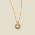A gold chain necklace with a circular pendant featuring light green baguette-cut stones around a hollow center, displayed against a solid light beige background.