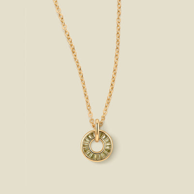 A gold chain necklace with a circular pendant featuring light green baguette-cut stones around a hollow center, displayed against a solid light beige background.