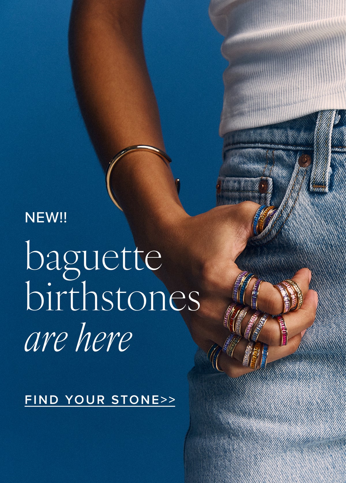 A person wearing multiple colorful baguette birthstone rings stacked on their fingers shown from the chest down in a white top and blue jeans against a solid blue background with promotional text.