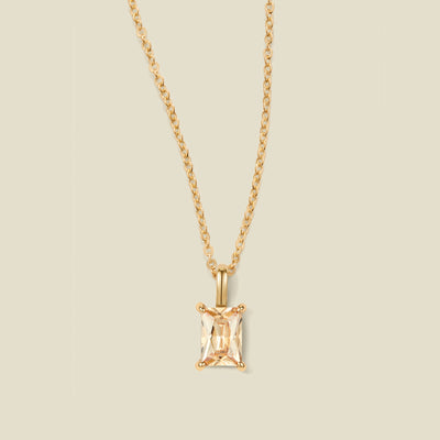 A gold chain necklace with a baguette-cut warm golden gemstone pendant in a gold prong setting, shown against a solid light beige background.