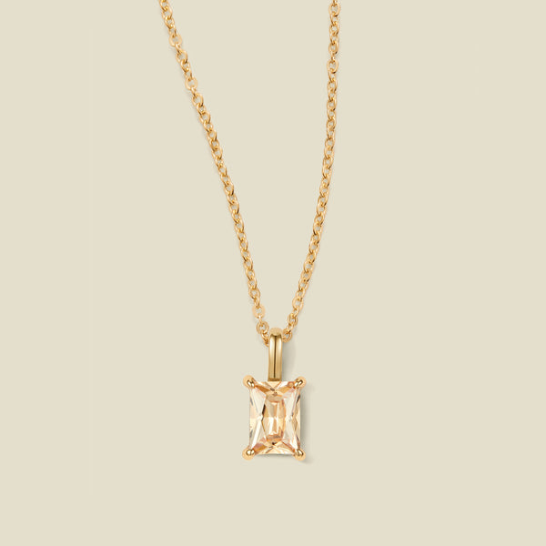 A gold chain necklace with a baguette-cut warm golden gemstone pendant in a gold prong setting, shown against a solid light beige background.