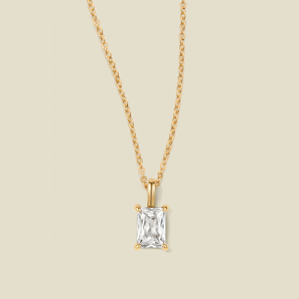 A gold chain necklace with a baguette-cut clear gemstone pendant, displayed against a solid light beige background.