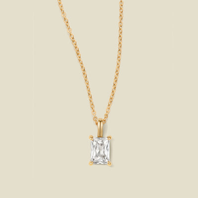 A gold chain necklace with a baguette-cut clear gemstone pendant, displayed against a solid light beige background.