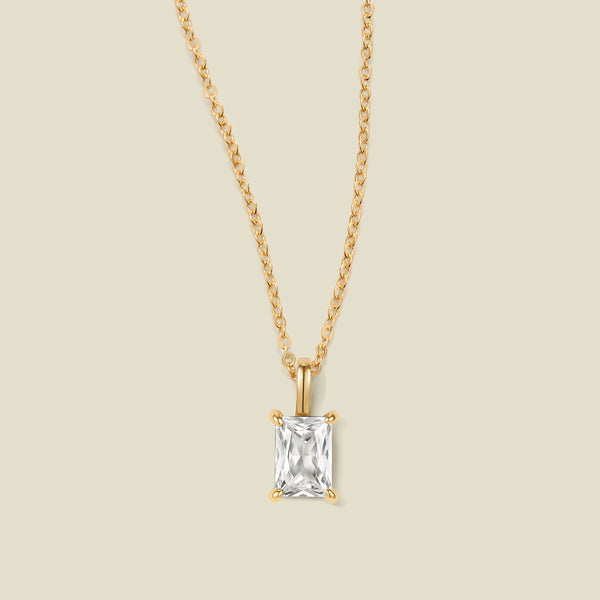 A gold chain necklace with a baguette-cut clear gemstone pendant, displayed against a solid light beige background.