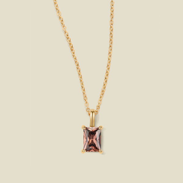 A gold chain necklace with a baguette-cut black/brown gemstone pendant, displayed against a solid light beige background.
