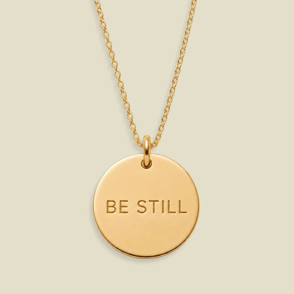 A gold disc pendant necklace engraved with the words 'BE STILL' in a clean sans-serif font, suspended from a delicate gold chain against a solid, warm beige background.