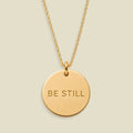 A gold disc pendant necklace engraved with the words 'BE STILL' in a clean sans-serif font, suspended from a delicate gold chain against a solid, warm beige background.