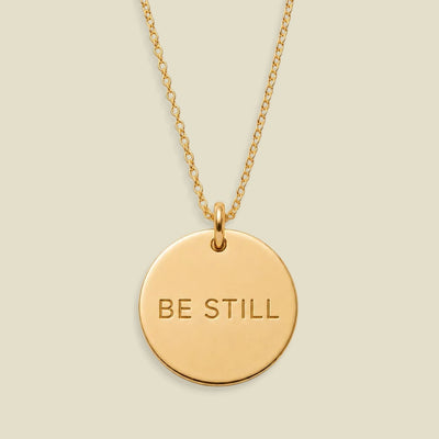 A gold disc pendant necklace engraved with the words 'BE STILL' in a clean sans-serif font, suspended from a delicate gold chain against a solid, warm beige background.