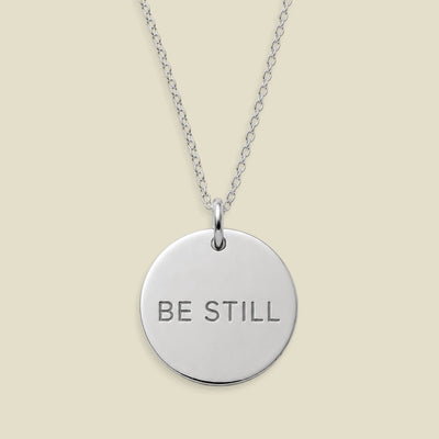 A silver round disc pendant necklace engraved with the words 'BE STILL' in capital letters, suspended from a delicate silver chain against a solid light beige background.