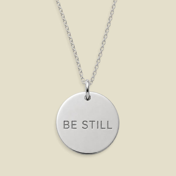 A silver round disc pendant necklace engraved with the words 'BE STILL' in capital letters, suspended from a delicate silver chain against a solid light beige background.