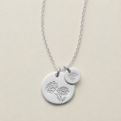 A silver necklace with two circular charms, one larger engraved with a floral design and one smaller disc with a different floral stamp, displayed on a light beige background.