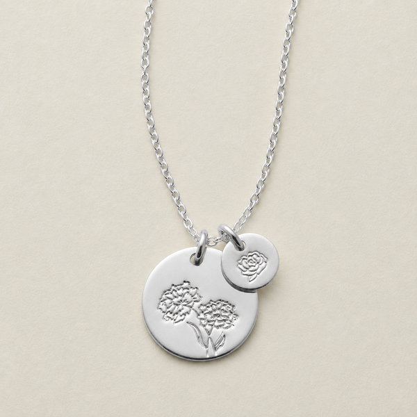A silver necklace with two circular charms, one larger engraved with a floral design and one smaller disc with a different floral stamp, displayed on a light beige background.