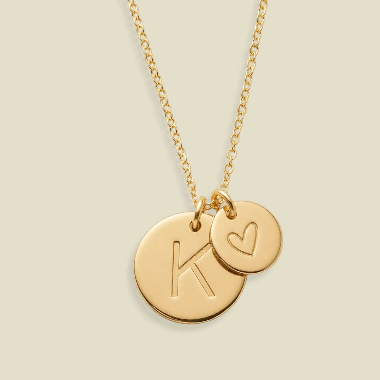 A gold chain necklace with two round pendant discs, one larger disc engraved with the letter 'K' and the other smaller disc with a heart, on a light beige background.