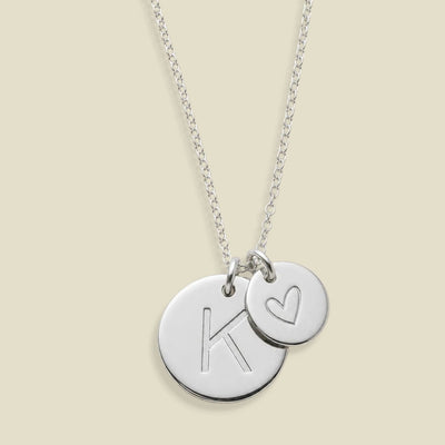 A silver chain necklace with two round disc charms, one bigger pendant engraved with a large letter K and the other smaller pendant with a heart, on a light beige background.