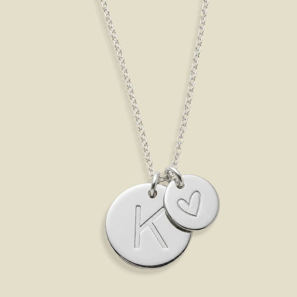 A silver chain necklace with two round disc charms, one bigger pendant engraved with a large letter K and the other smaller pendant with a heart, on a light beige background.