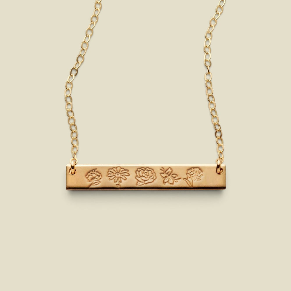 A gold bar necklace with a flat horizontal bar engraved with five different flower motifs, suspended from a delicate gold chain against a solid light beige background.