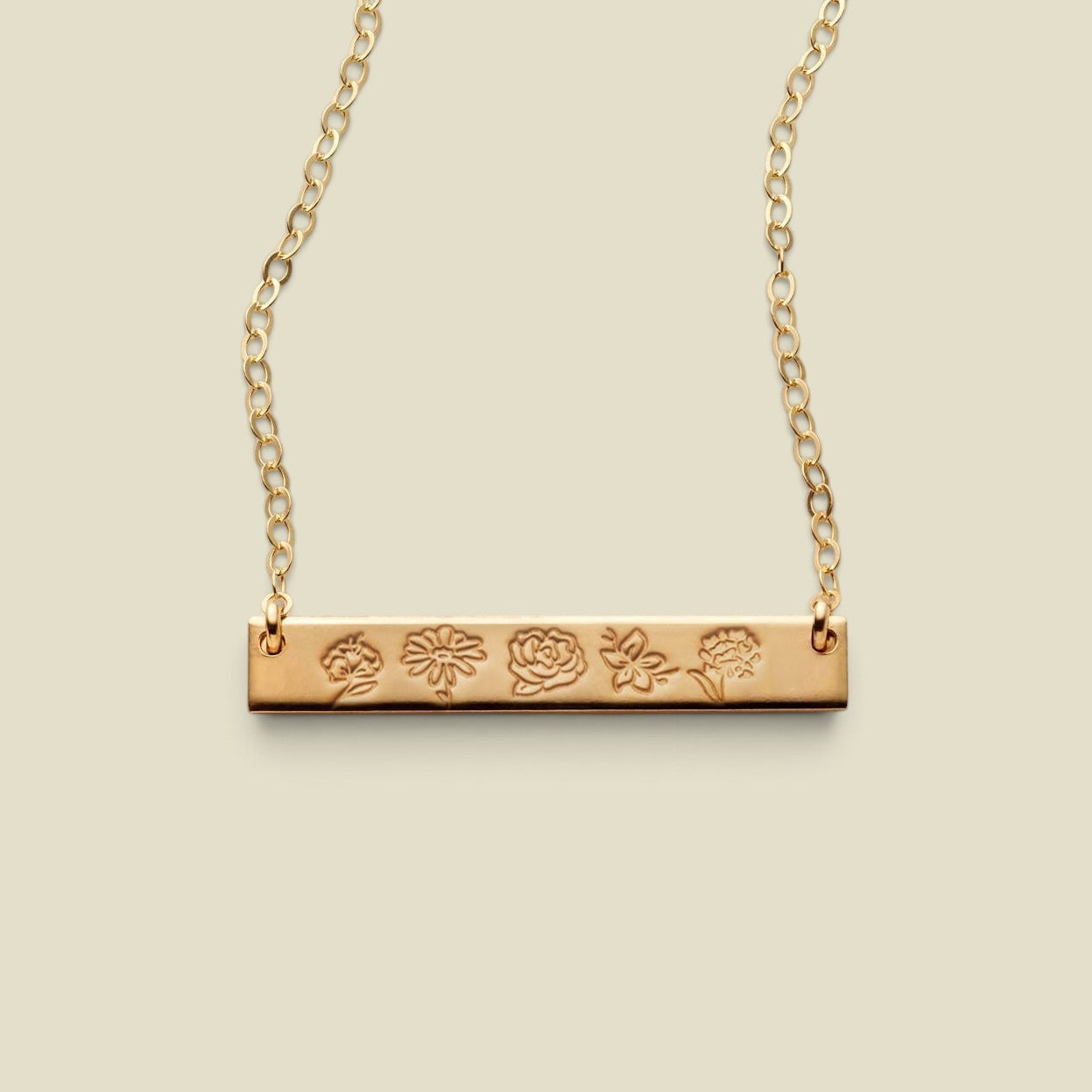 A gold bar necklace with a flat horizontal bar engraved with five different flower motifs, suspended from a delicate gold chain against a solid light beige background.
