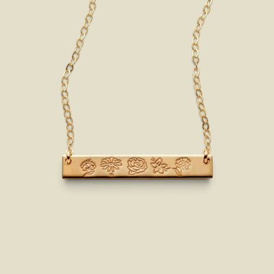 A gold bar necklace with a flat horizontal bar engraved with five different flower motifs, suspended from a delicate gold chain against a solid light beige background.