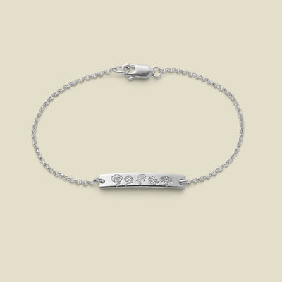 A delicate silver chain bracelet with a horizontal bar, engraved with different birth flowers, set against a soft beige background.
