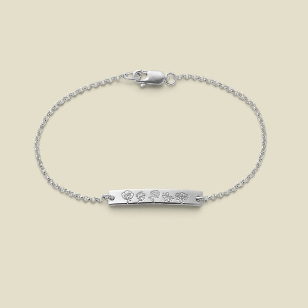 A delicate silver chain bracelet with a horizontal bar, engraved with different birth flowers, set against a soft beige background.