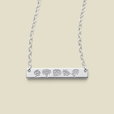 A silver bar necklace with five small engraved floral motifs, suspended from a delicate silver chain against a solid beige background.