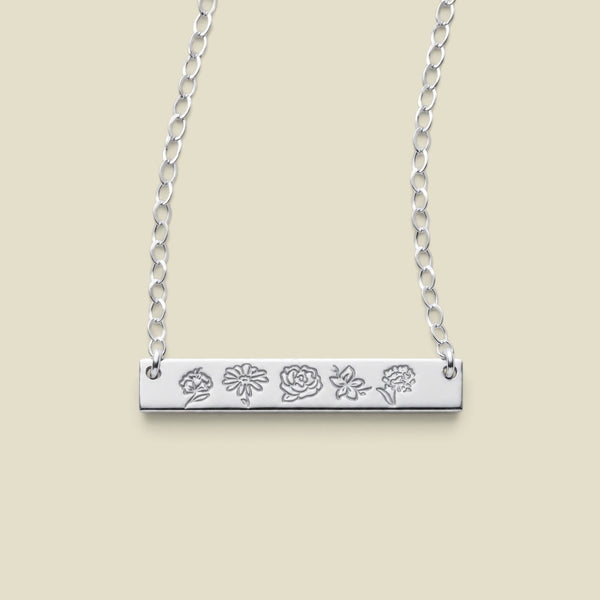 A silver bar necklace with five small engraved floral motifs, suspended from a delicate silver chain against a solid beige background.