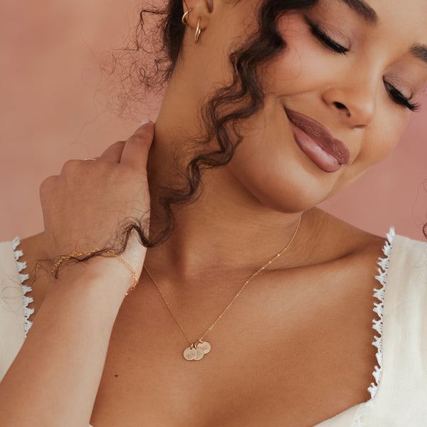 A close-up lifestyle shot of a woman wearing a delicate gold necklace with three round discs, each with different birth flowers engraved, styled with a matching gold bracelet. She is wearing a white top with scalloped edges against a soft, solid dusty pink background.