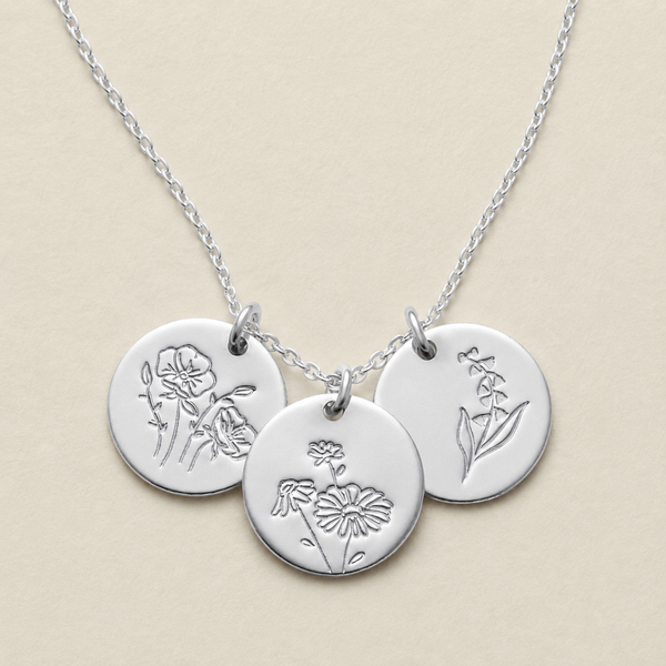 A silver chain necklace with three round engraved disc pendants featuring delicate floral designs, displayed on a warm beige background.