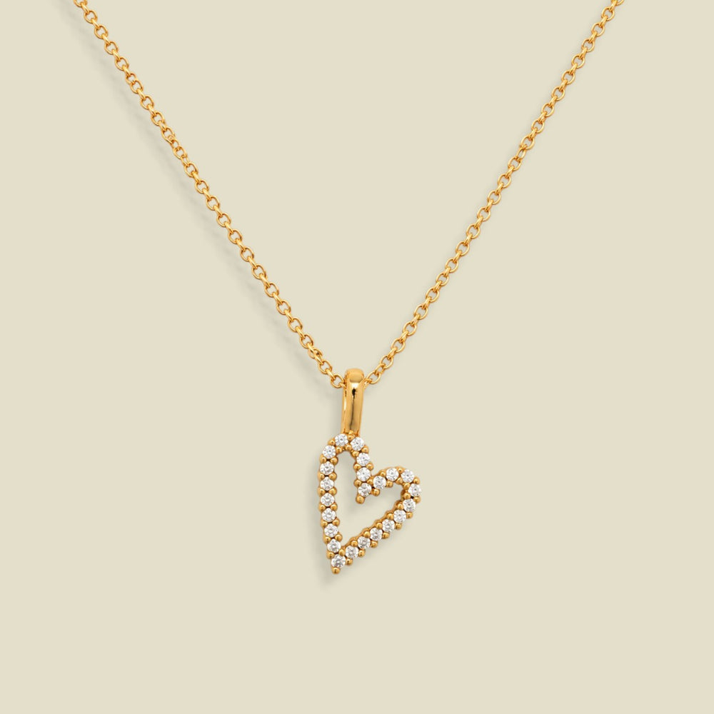 A gold chain necklace with a heart-shaped pendant outlined in small clear stones, displayed on a solid light beige background.