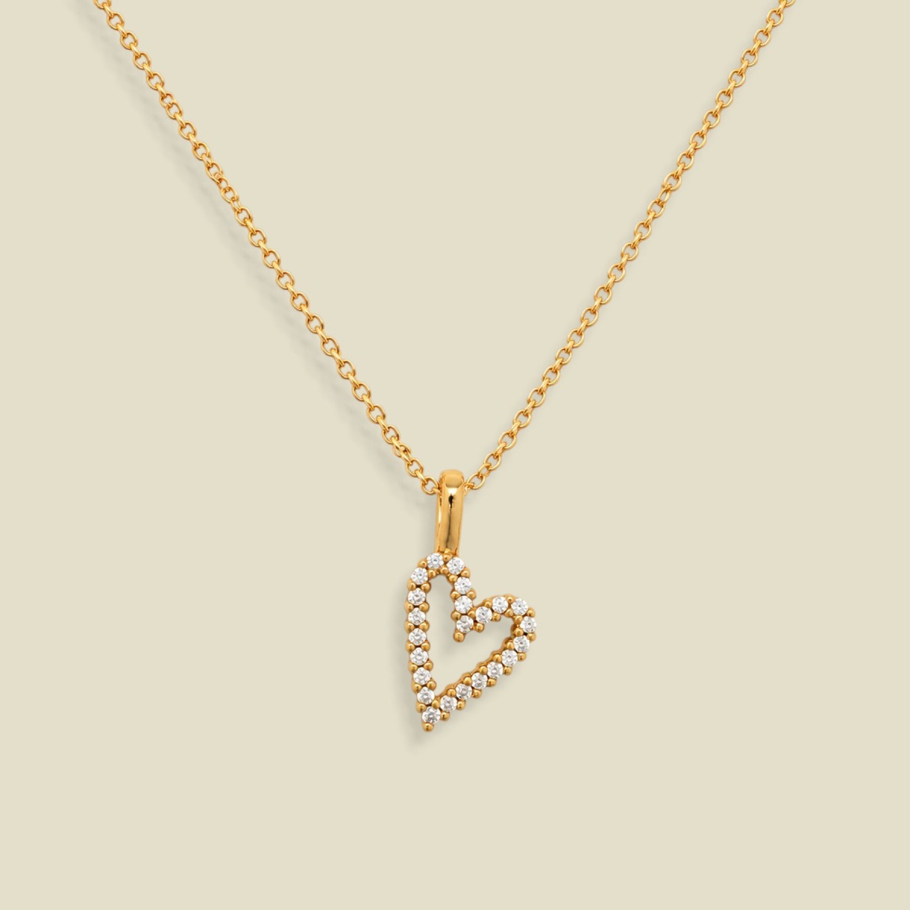 A gold chain necklace with a heart-shaped pendant outlined in small clear stones, displayed on a solid light beige background.