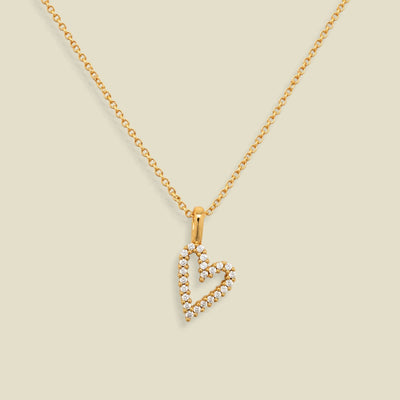 A gold chain necklace with a heart-shaped pendant outlined in small clear stones, displayed on a solid light beige background.