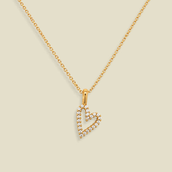 A gold chain necklace with a heart-shaped pendant outlined in small clear stones, displayed on a solid light beige background.