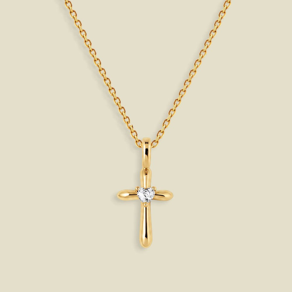 A gold cross pendant necklace with a small clear heart-shaped gemstone at the center, displayed on a light beige background.
