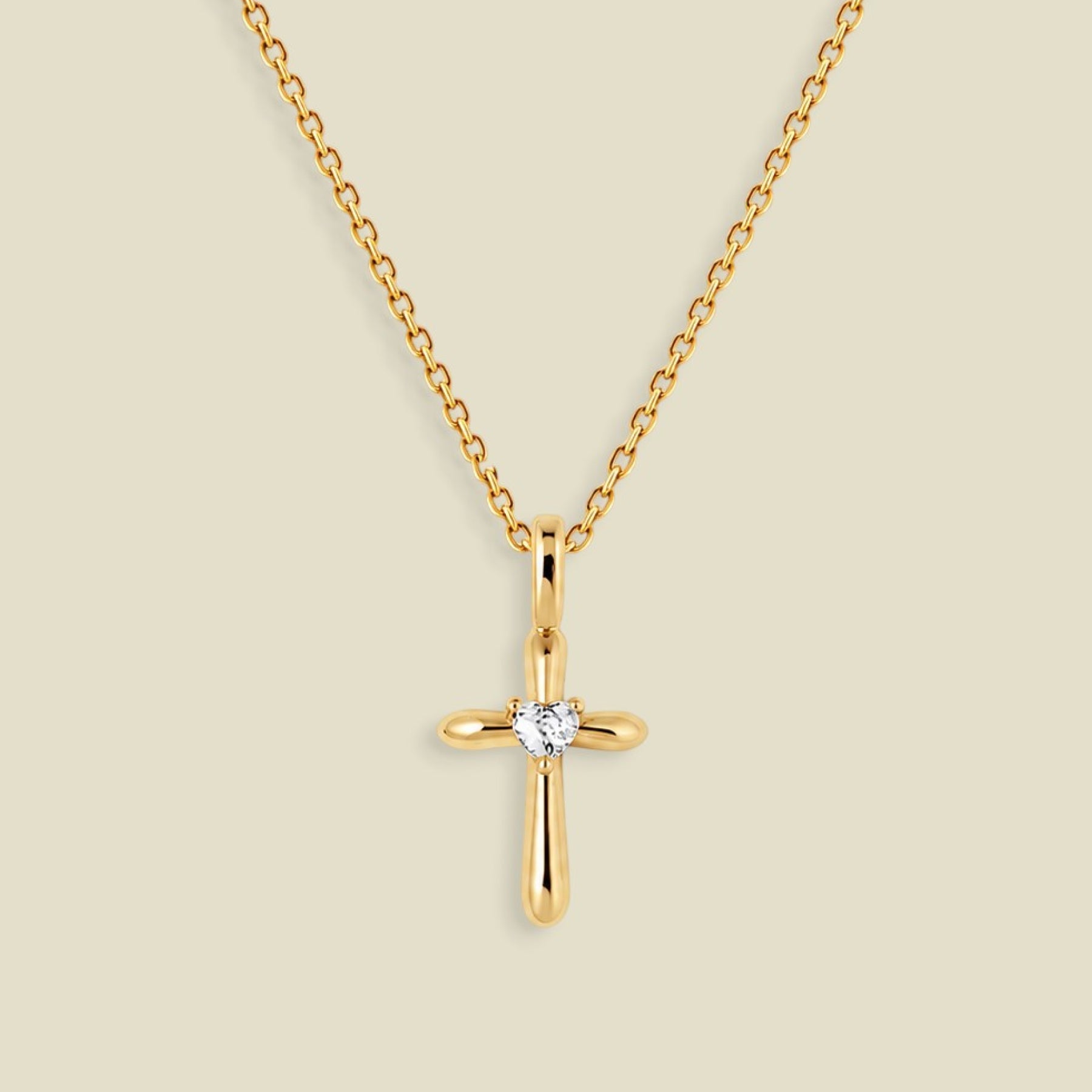 A gold cross pendant necklace with a small clear heart-shaped gemstone at the center, displayed on a light beige background.