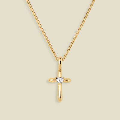 A gold cross pendant necklace with a small clear heart-shaped gemstone at the center, displayed on a light beige background.