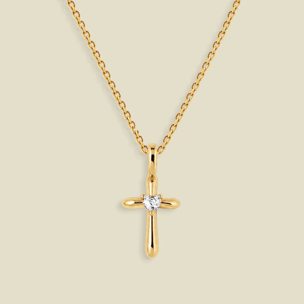 A gold cross pendant necklace with a small clear heart-shaped gemstone at the center, displayed on a light beige background.