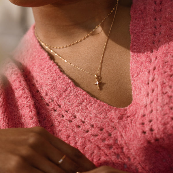 A close-up lifestyle shot of a person wearing a delicate gold cross pendant necklace with a cz heart, layered over a gold chain, paired with a pink knit sweater.