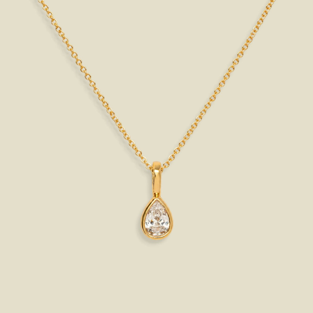 A gold chain necklace with a teardrop-shaped clear gemstone pendant in a gold bezel setting, displayed against a solid light beige background.