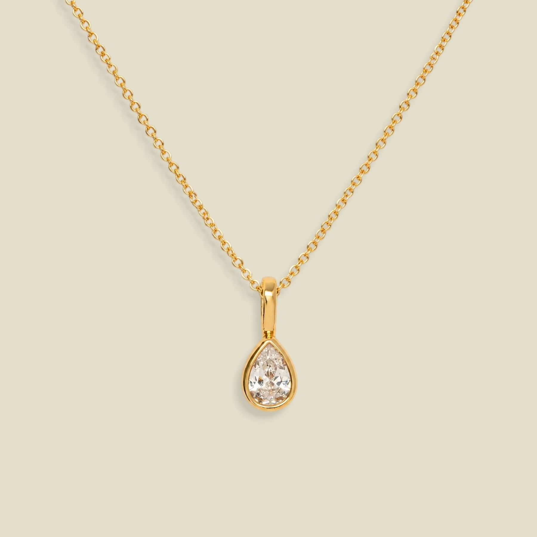 A gold chain necklace with a teardrop-shaped clear gemstone pendant in a gold bezel setting, displayed against a solid light beige background.