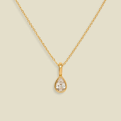 A gold chain necklace with a teardrop-shaped clear gemstone pendant in a gold bezel setting, displayed against a solid light beige background.