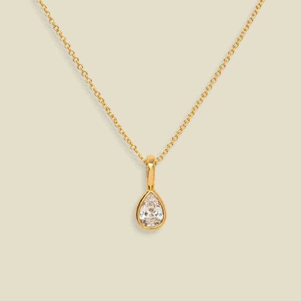A gold chain necklace with a teardrop-shaped clear gemstone pendant in a gold bezel setting, displayed against a solid light beige background.