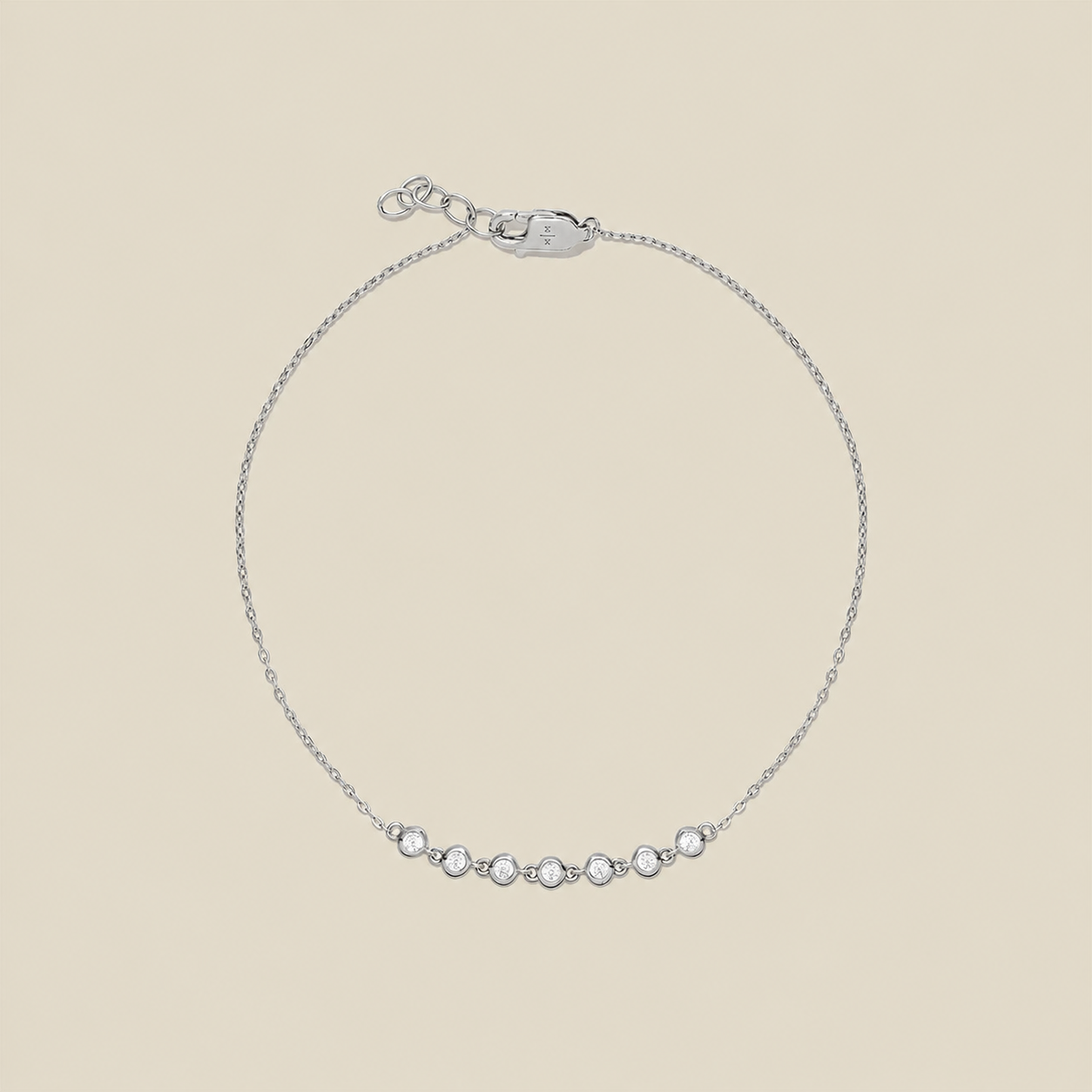 A delicate silver chain bracelet with a row of 7 small clear bezel-set round stones across the center, displayed on a solid light beige background.