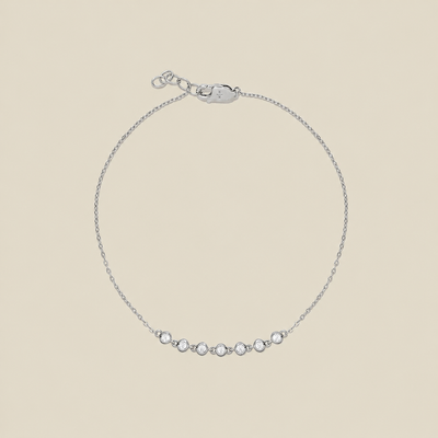A delicate silver chain bracelet with a row of 7 small clear bezel-set round stones across the center, displayed on a solid light beige background.