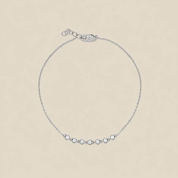 A delicate silver chain bracelet with a row of 7 small clear bezel-set round stones across the center, displayed on a solid light beige background.
