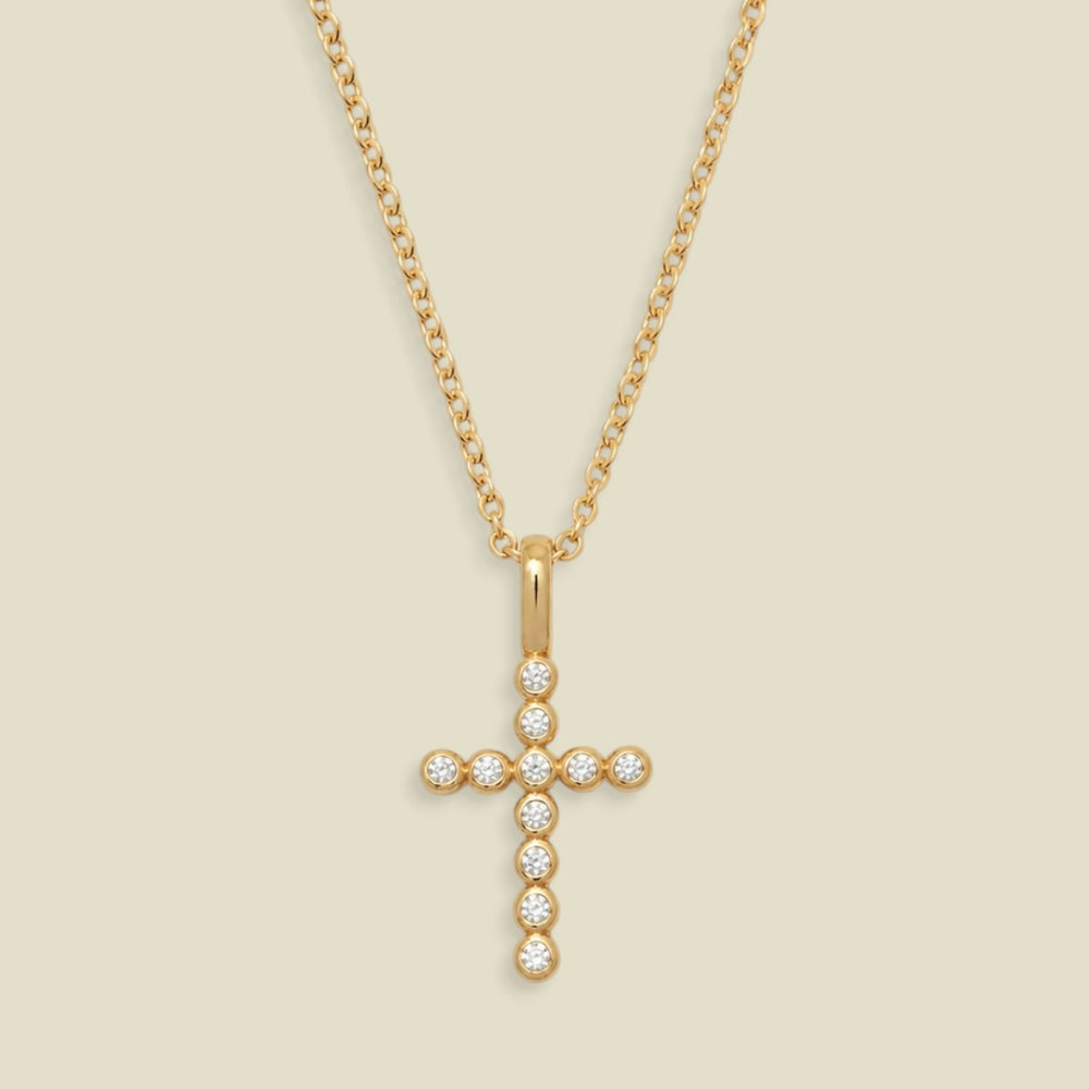 A gold cross pendant necklace adorned with small clear round bezel set gemstones, suspended from a delicate gold chain against a light beige background.