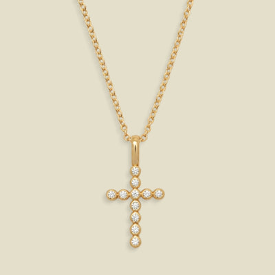A gold cross pendant necklace adorned with small clear round bezel set gemstones, suspended from a delicate gold chain against a light beige background.